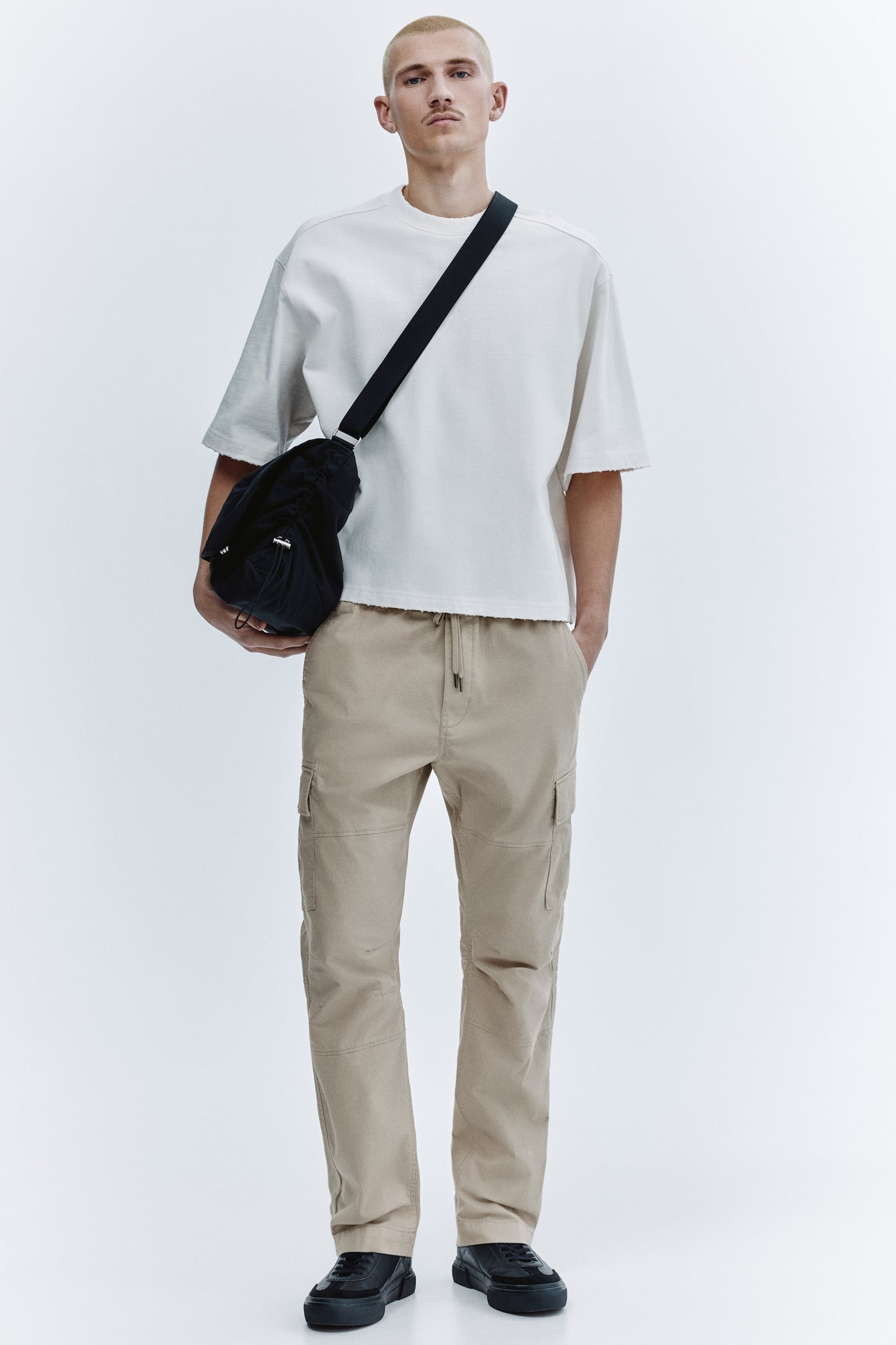 Person wearing a white t-shirt, beige cargo pants, and black shoes with a black crossbody bag on a white background.