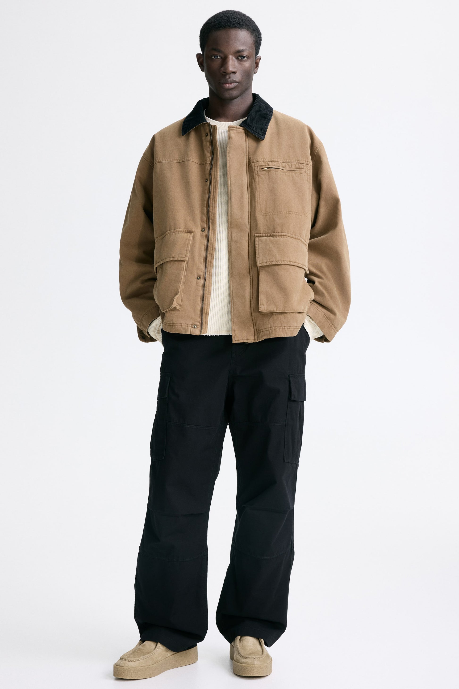 Man wearing a beige jacket with black collar and black pants on a white background