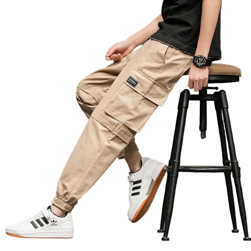 Person wearing beige cargo pants and white Adidas sneakers sitting on a black stool.
